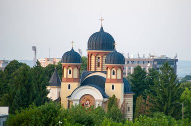 Girişin üzerinde mozaiği olan üç kubbeli bir kilise ağaçların ve şehir binalarının arasında duruyor..