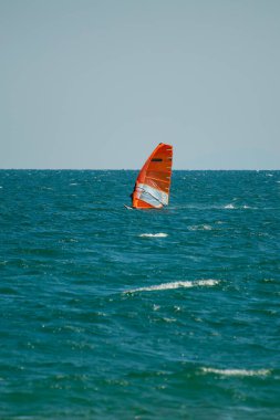 Bir rüzgar sörfçüsü berrak bir gökyüzünün altında canlı turuncu bir yelkenle okyanusta süzülüyor..