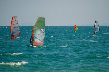 Renkli yelkenli bir grup rüzgar sörfçüsü, Bulgaristan 'ın ufukta görünen uzak adaları tarafından çerçevelenmiş berrak bir gökyüzü altında dalgalı mavi-yeşil sularda süzülüyor.
