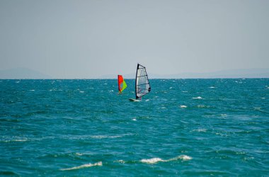 İki rüzgar sörfçüsü açık bir gökyüzü altında mavi-yeşil dalgaları kesiyor, yelkenleri uzak ufka karşı cesurca tezat oluşturuyor..