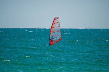 Şeffaf turuncu yelkenli bir rüzgar sörfçüsü açık bir gökyüzünün altında açık mavi-yeşil dalgalar üzerinde süzülür ve rüzgarın gücünü kucaklar..