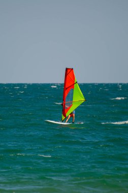 Canlı kırmızı, yeşil ve sarı yelkenli bir rüzgar sörfçüsü, Bulgaristan 'da açık mavi bir gökyüzü altında turkuaz okyanus dalgaları arasında süzülüyor