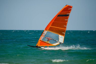 Bir rüzgar sörfçüsü berrak bir gökyüzünün altında canlı turuncu bir yelkenle okyanusta süzülüyor..