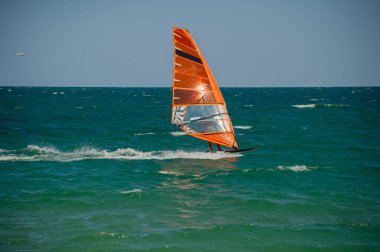 Şeffaf turuncu yelkenli bir rüzgar sörfçüsü açık bir gökyüzünün altında açık mavi-yeşil dalgalar üzerinde süzülür ve rüzgarın gücünü kucaklar..