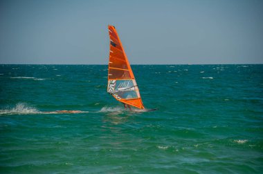 Şeffaf turuncu yelkenli bir rüzgar sörfçüsü açık bir gökyüzünün altında açık mavi-yeşil dalgalar üzerinde süzülür ve rüzgarın gücünü kucaklar..