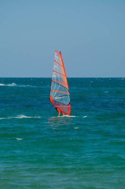 Şeffaf turuncu yelkenli bir rüzgar sörfçüsü açık bir gökyüzünün altında açık mavi-yeşil dalgalar üzerinde süzülür ve rüzgarın gücünü kucaklar..
