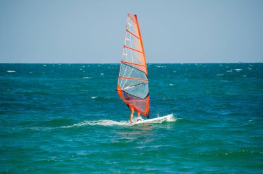 Şeffaf turuncu yelkenli bir rüzgar sörfçüsü açık bir gökyüzünün altında açık mavi-yeşil dalgalar üzerinde süzülür ve rüzgarın gücünü kucaklar..
