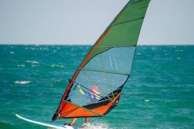 Rüzgâr sörfçüsü dalgalı okyanus dalgalarını hızlı bir şekilde geçiyor. Canlı yeşil, turuncu ve mavi yelkenli ile. Rüzgarlı, güneşli bir günde..