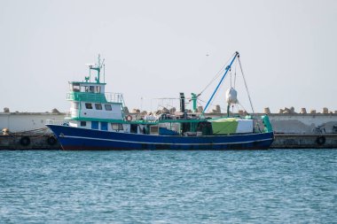  Orta büyüklükte bir mavi, beyaz ve yeşil balıkçı teknesi, arka planda tetrapodlar bulunan beton bir iskelenin yanında demirli..