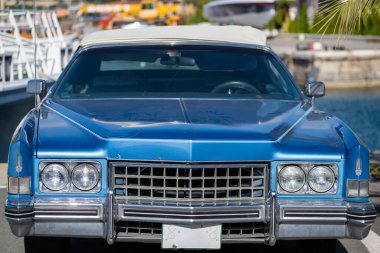 Beyaz kapaklı, klasik, parlak mavi bir Cadillac arka planda bulanık tekneler ve sarı ekipmanlarla bir rıhtımın yakınına park edilmiş..