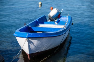  Dış motorlu küçük beyaz ve mavi bir balıkçı teknesi, arka planda iki şamandıranın yüzdüğü berrak, durgun mavi bir suya bağlanmış durumda..