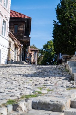  Nesebar 'ın tarihi bir kasabasında tepeden tırnağa uzanan bir kaldırım taşı caddesi kırmızı çatılı binalar ve güneşli bir gökyüzünün altında yapraklı ağaçlarla kaplı..