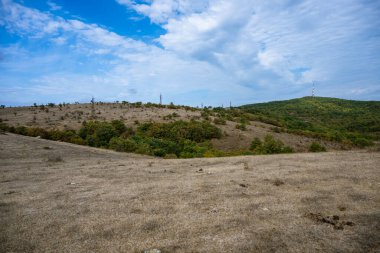Haberleşme kuleleri ormanlık tepelerin ve kuru otlakların üzerinde parçalı bulutlu bir gökyüzünün altında yükseliyor.,