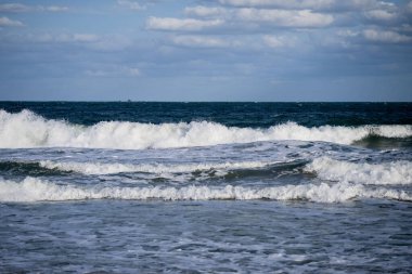 Dalgalar, parçalı bulutlu bir gökyüzünün altında denizin ham enerjisini ve ritmini yakalayarak derin mavi denizde çarpışıp yuvarlanırlar..