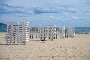 Beyaz şezlonglar, kumlu bir sahilde, yumuşak, bulutlu bir gökyüzünün altında güneşlenenlerin dönüşünü bekliyor..