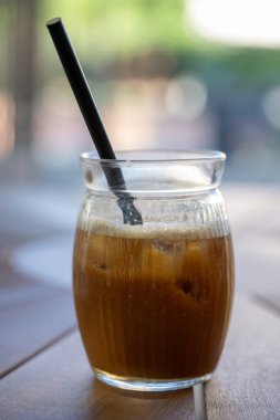 Köpüklü buzlu bir bardak buzlu kahve ve siyah bir pipet güneşli bir kafede, ahşap bir masada oturur.
