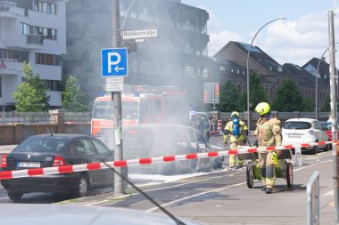 Tam teçhizatlı itfaiyeciler bir araba yangınını beyaz köpükle ya da aracın etrafında uçuşan dumanla söndürüyorlar. Yanan arabanın arkasında bir itfaiye aracı görünüyor ve yakınlarda bir polis cipi var. Sahne kırmızı ve beyaz çizgili bariyerler tarafından kordon altına alındı.