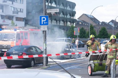 Tam teçhizatlı itfaiyeciler bir araba yangınını beyaz köpükle ya da aracın etrafında uçuşan dumanla söndürüyorlar. Yanan arabanın arkasında bir itfaiye aracı görünüyor ve yakınlarda bir polis cipi var. Sahne kırmızı ve beyaz çizgili bariyerler tarafından kordon altına alındı.