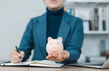 Piggy Bank ve Finansal Planlama Konsepti. İş kadını elinde dizüstü bilgisayarla pembe bir kumbara tutuyor. Finansal planlama, tasarruf, yatırım ve para yönetimini sembolize ediyor..