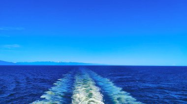 Canlı bir mavi okyanus berrak bir gökyüzünün altında uzanır, köpüklü bir teknenin uyanışıyla. Uzak dağlar ufuk çizgisinde.