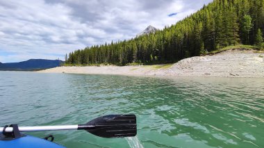 Bir teknenin manzarası, açık turkuaz dağ sularını, yemyeşil ormanlarla ve bulutlu gökyüzünün altındaki görkemli tepelerle çevrili bir kürekle gösterir. Açık hava macerası için idealdir.
