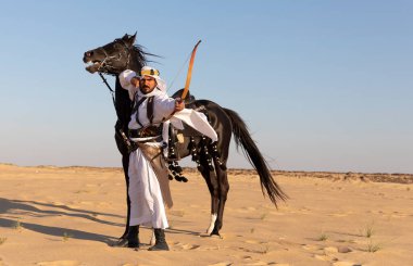 Siyah bir aygırla çölde geleneksel Suudi Arabistan kıyafetleri giyen bir adam.