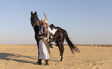 Siyah bir aygırla çölde geleneksel Suudi Arabistan kıyafetleri giyen bir adam.