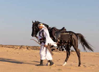 Siyah bir aygırla çölde geleneksel Suudi Arabistan kıyafetleri giyen bir adam.