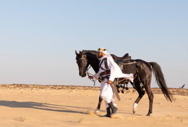 Siyah bir aygırla çölde geleneksel Suudi Arabistan kıyafetleri giyen bir adam.