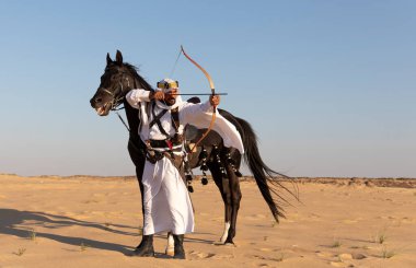 Siyah bir aygırla çölde geleneksel Suudi Arabistan kıyafetleri giyen bir adam.