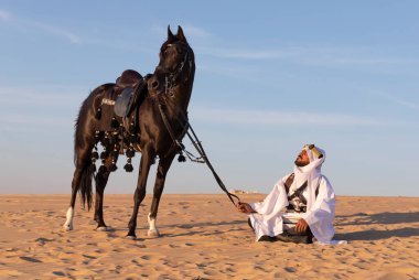 Siyah bir aygırla çölde geleneksel Suudi Arabistan kıyafetleri giyen bir adam.