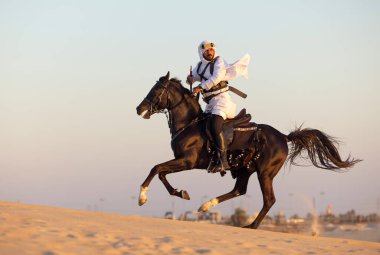 Siyah bir aygırla çölde geleneksel Suudi Arabistan kıyafetleri giyen bir adam.