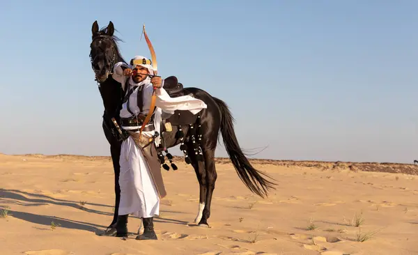 Man in traditional Saudi Arabian clothing in a desert with a black stallion