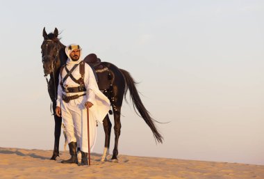 Siyah bir aygırla çölde geleneksel Suudi Arabistan kıyafetleri giyen bir adam.