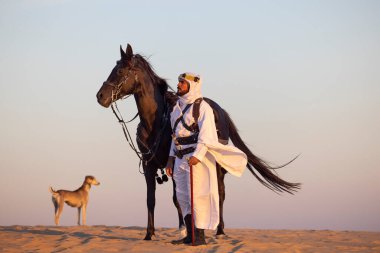 Siyah bir aygırla çölde geleneksel Suudi Arabistan kıyafetleri giyen bir adam.