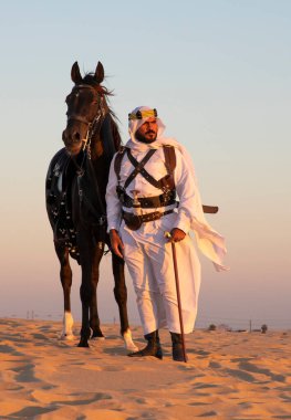 Siyah bir aygırla çölde geleneksel Suudi Arabistan kıyafetleri giyen bir adam.