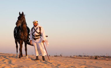 Siyah bir aygırla çölde geleneksel Suudi Arabistan kıyafetleri giyen bir adam.