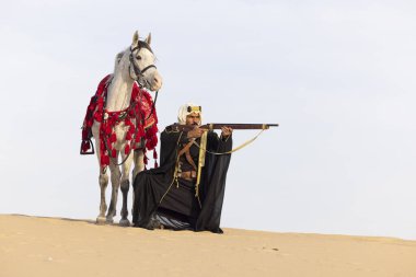 Suudi adam, geleneksel kıyafetleriyle çölde beyaz aygırıyla tüfeğini doğrultuyor.