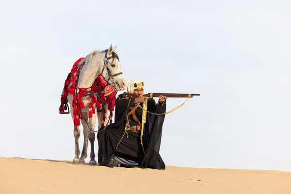 Saudi man in traditional clothing with his white stallion in a desert, aiming a rifle