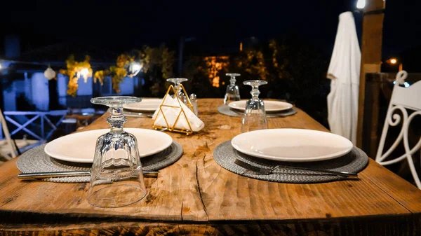Empty table decorated with glasses, cutlery, dishes and place mats on the terrace on a summer evening. An empty decorated table ready to serve people.