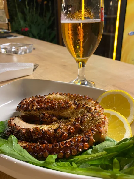 Close up view of a grilled octopus with sliced lemon, arugula served on plate with cold beer. greek style