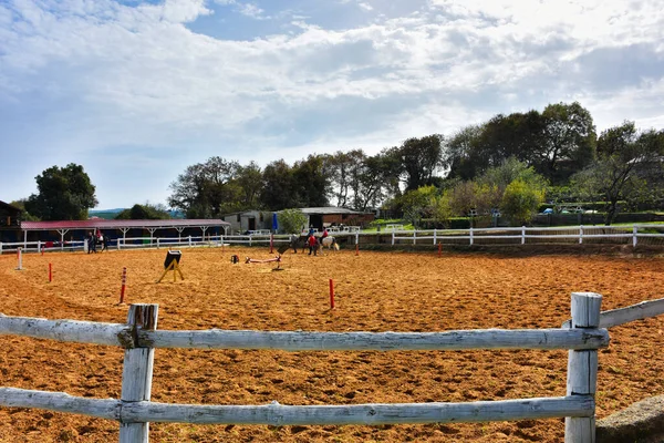 Riding training equipments, a horse, people silhouettes  on training track in a horse farm with trees and clouds sky view. Wide angle view of equestrian training track in a horse farm.