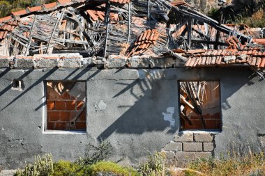 Front view of an abandoned concrete house in the countryside. Ruined wood and brick roof. frameless windows and swaths of dry and green grass