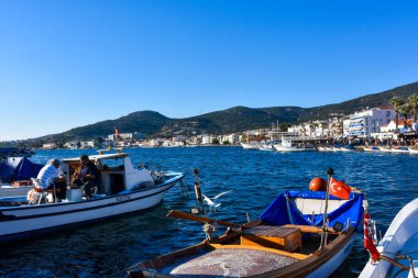 Izmir, Foca Turkey. 07.06.2021 Turkish flagged fishing boats along the coastline of Foa resort, two old people sitting on the boat, hotels and restaurants on the opposite shore. Touristic Aegean town