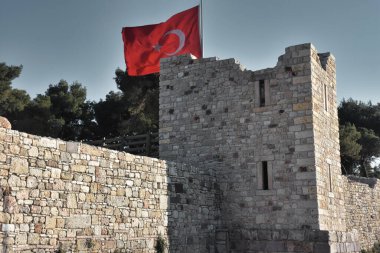 A large Turkish flag, trees and a clear sky behind the ruins of a destroyed old medieval castle. Izmir Foca