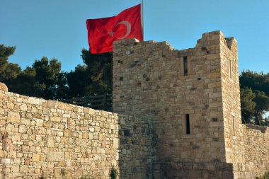 A large Turkish flag, trees and a clear sky behind the ruins of a destroyed old medieval castle. Izmir Foca