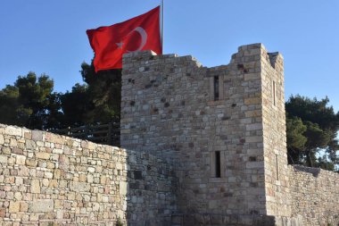 A large Turkish flag, trees and a clear sky behind the ruins of a destroyed old medieval castle. Izmir Foca