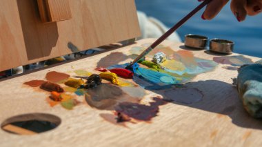 Genç bayan ressam sehpanın önünde duruyor, yağlı boyaları fırçasıyla karıştırıyor. Plein hava boyası.