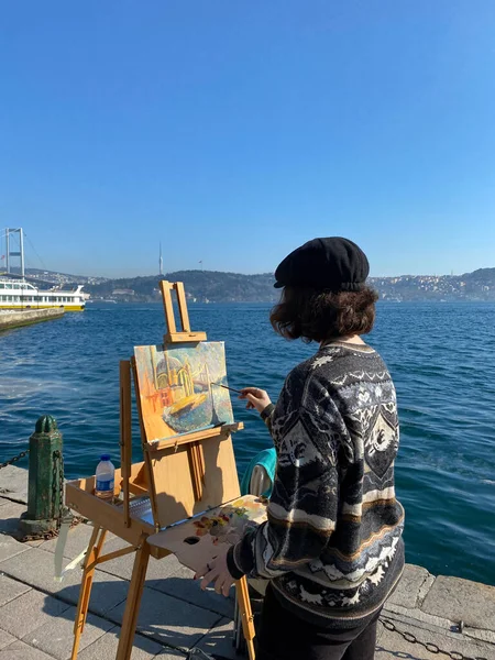 The young plein air painter artist look at the Ortakoy Mosque on the shore of Ortakoy and paints it front of her easel. Besiktas, Istanbul, Turkey