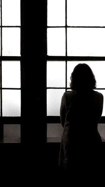 Eski tarz trençkot giymiş genç bir kadın pencerenin kenarında durur arkasını döner ve pencereden dışarı bakar..